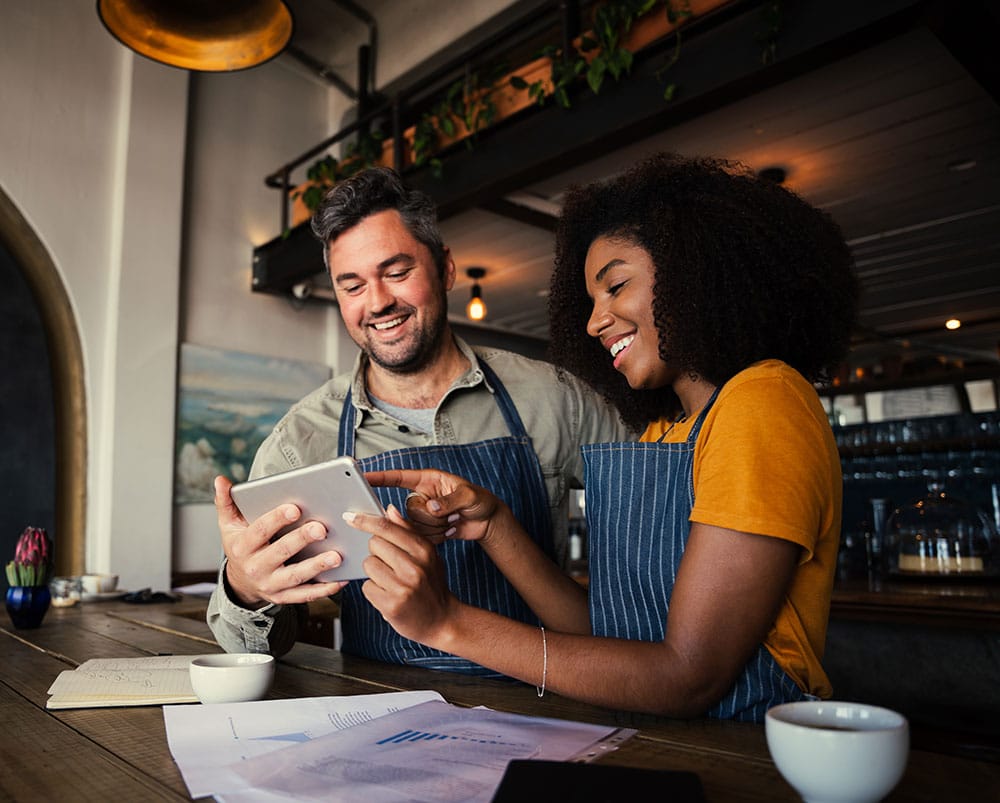 Manager and waitress laughing at designs on digital tablet standing with hot coffee in cafe